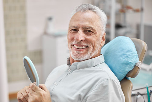 The image features a smiling man with gray hair, seated in a dental chair and holding a mirror, dressed in a white shirt and blue jeans. He is in a dental office setting, with dental equipment visible behind him.
